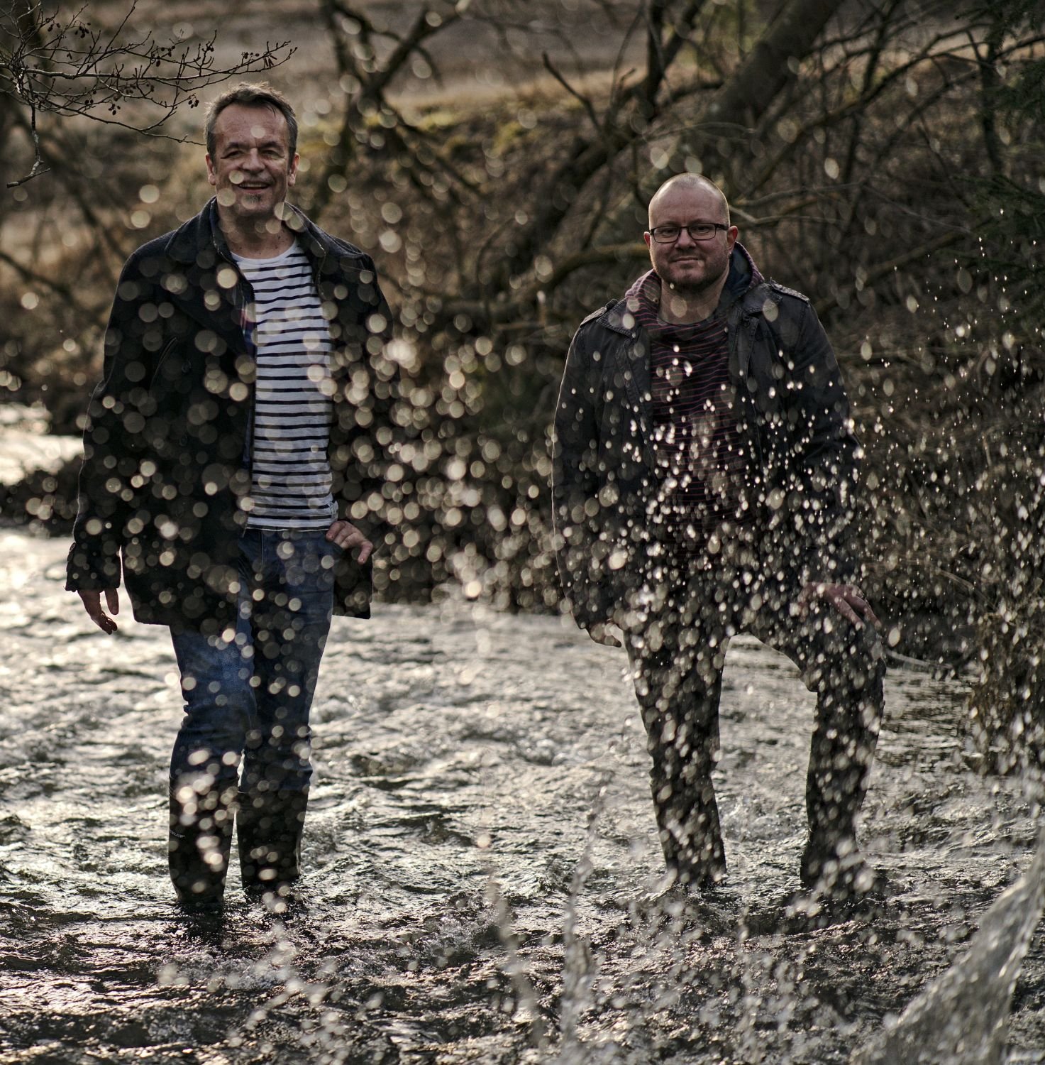 Pascal Nicolas und Dirk Wilbert stehen in Gummistiefeln in einem Bach, Wassertropfen spritzen im Gegenlicht.